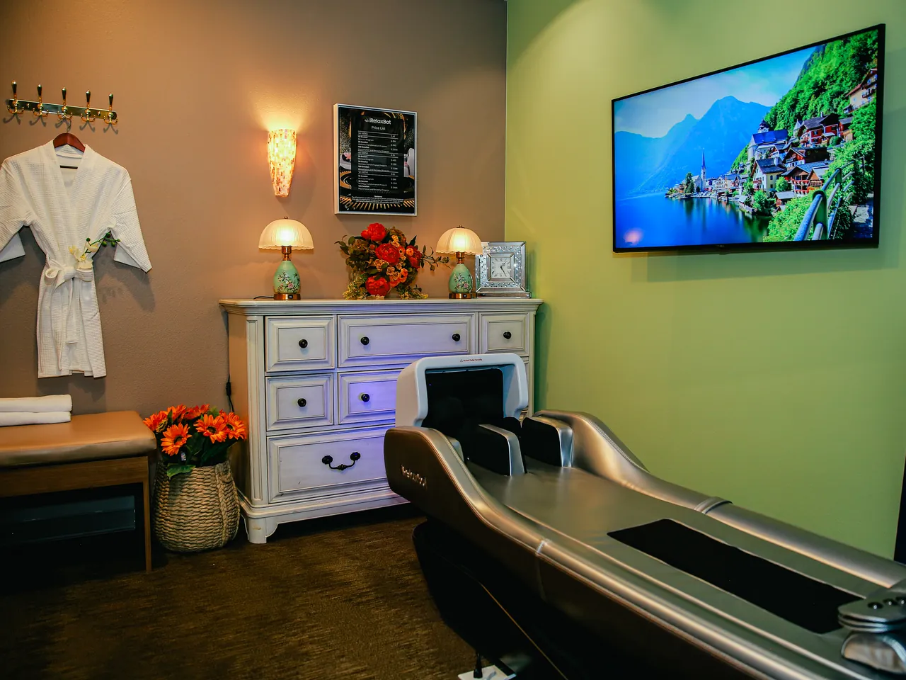 The iRelaxbot robotic massage bed in its bright private room — silver table, wall TV showing an alpine lake, white dresser with lamps and flowers, and a hanging spa robe